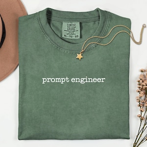 May include: A folded, olive green t-shirt with the words "prompt engineer" in white. A gold-colored necklace with a star pendant rests on the shirt. A brown hat is visible in the upper left corner.