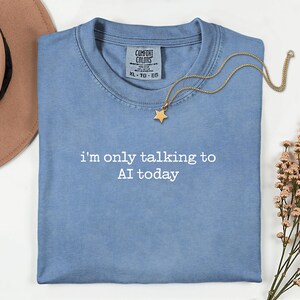 May include: A pale blue t-shirt with the white text "i'm only talking to AI today". A gold star pendant necklace rests on the shirt. The shirt is folded on a white surface.