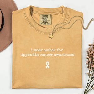 May include: A mustard yellow short-sleeve t-shirt with the white text "i wear amber for appendix cancer awareness" and a small white awareness ribbon icon printed on the front. The shirt features a Comfort Colors brand label at the inner neckline.