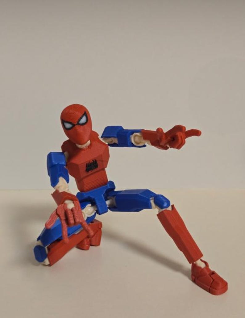 May include: A red, blue, and white articulated Spider-Man action figure in a crouching pose, pointing with one hand. The figure has a red mask with white eyes, a blue torso and legs, and red arms and feet. A black spider emblem is on the chest.
