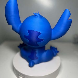 Może przedstawiać: Niebieska figurka Stitcha z filmu animowanego Lilo & Stitch. Figurka siedzi na białej platformie. Ma duże uszy, okrągły korpus i ciemnoniebieską plamę na plecach.