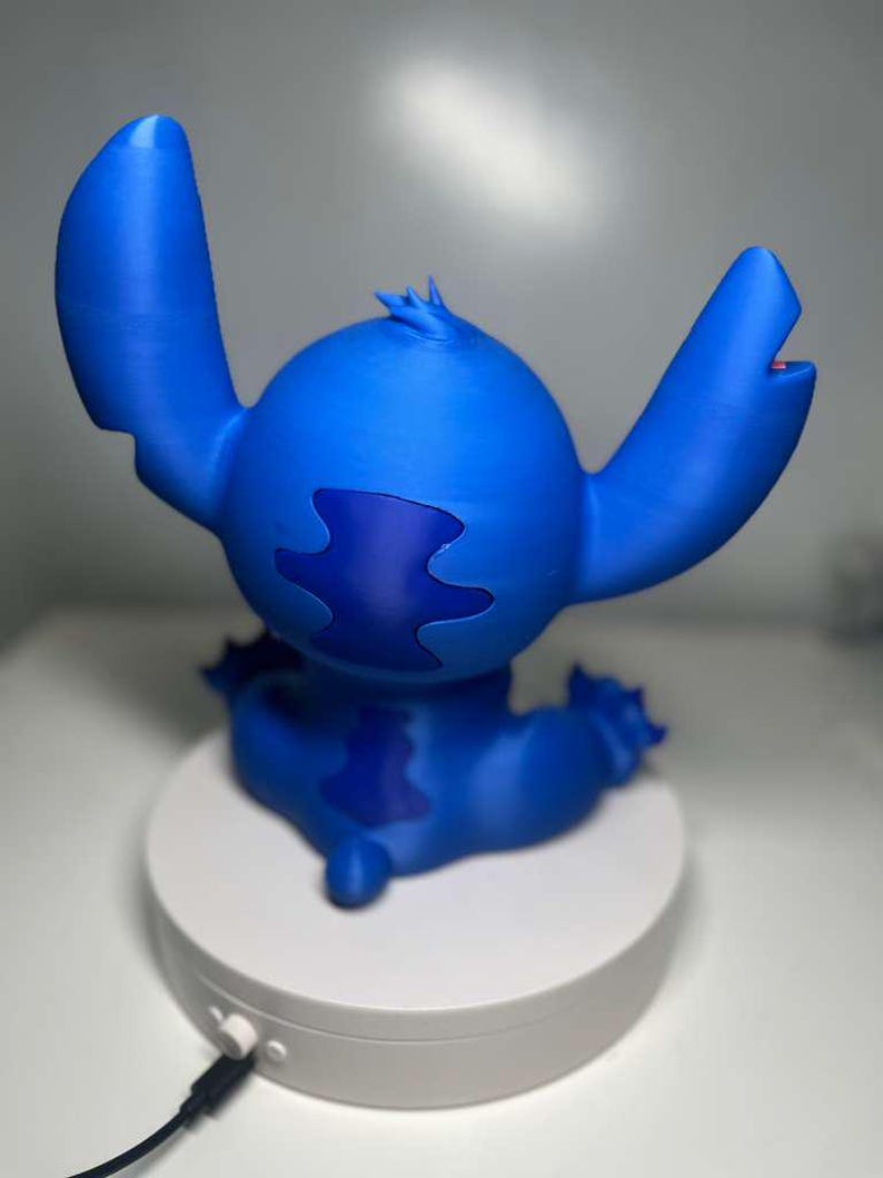 Może przedstawiać: Niebieska figurka Stitcha z filmu animowanego Lilo & Stitch. Figurka siedzi na białej platformie. Ma duże uszy, okrągły korpus i ciemnoniebieską plamę na plecach.