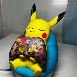 May include: A yellow Pikachu figurine with closed eyes, red cheeks, and black-tipped ears, holding a patterned video game controller. The controller features a floral and tiger design. The figurine is seated on a blue cushion.