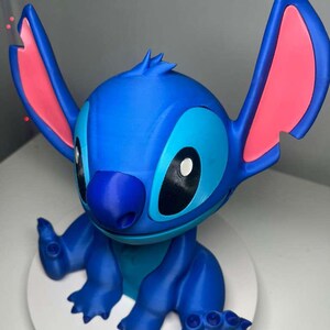 Może przedstawiać: Niebieska figurka postaci Stitch z filmu Disneya Lilo & Stitch. Figurka ma duże uszy z r&oacute;żowymi wnętrzami, ciemnoniebieski nos i duże czarne oczy. Znajduje się na białej, okrągłej podstawie.