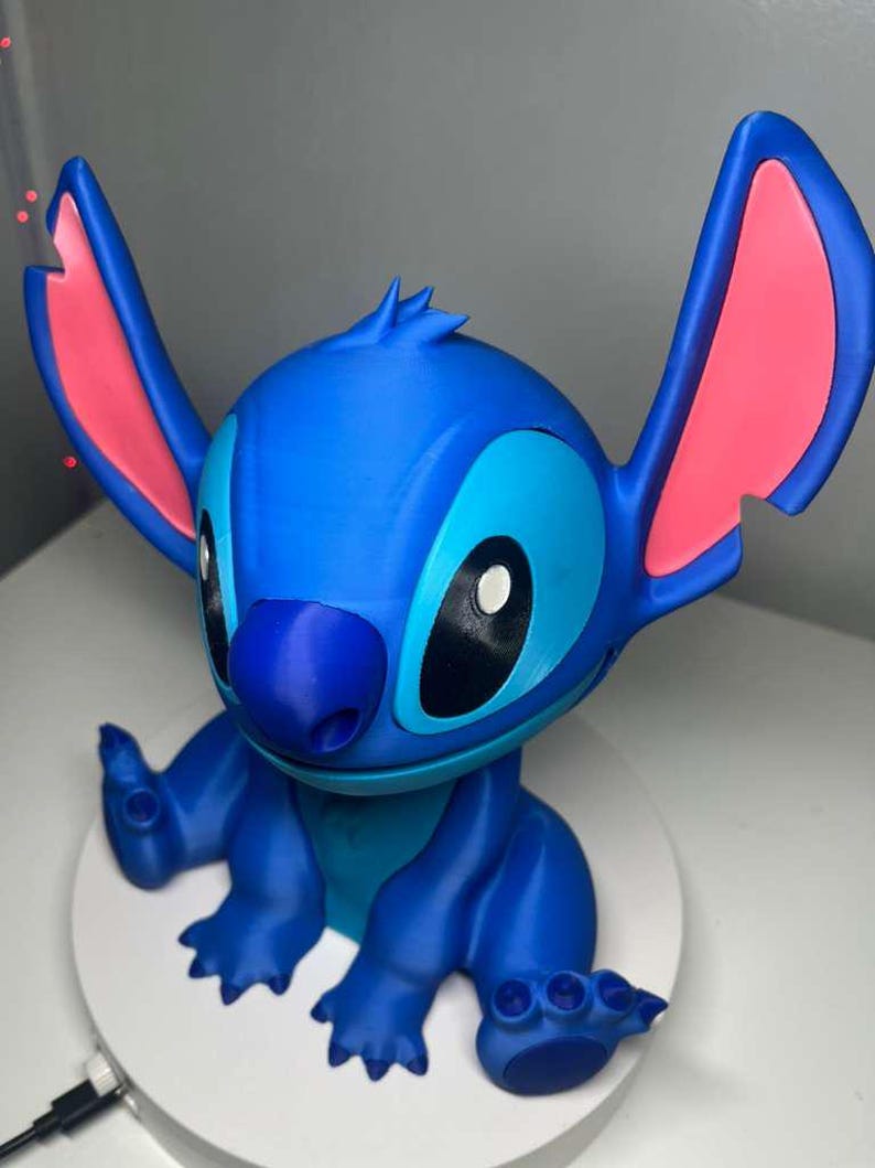Może przedstawiać: Niebieska figurka postaci Stitch z filmu Disneya Lilo & Stitch. Figurka ma duże uszy z r&oacute;żowymi wnętrzami, ciemnoniebieski nos i duże czarne oczy. Znajduje się na białej, okrągłej podstawie.
