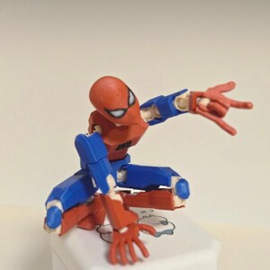 May include: A red, blue, and white Spider-Man action figure in a crouching pose. The figure is articulated, with visible joints and a detailed design. The figure is on a white surface.