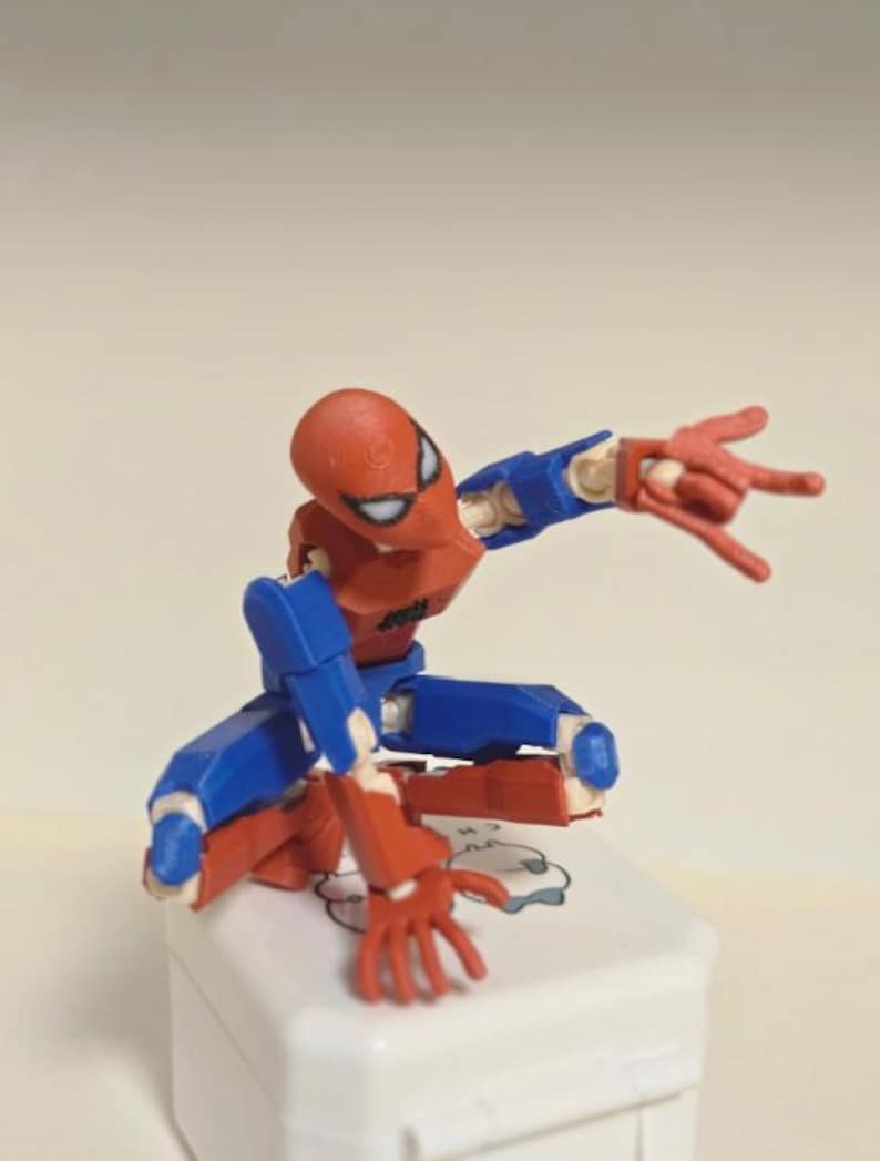 May include: A red, blue, and white Spider-Man action figure in a crouching pose. The figure is articulated, with visible joints and a detailed design. The figure is on a white surface.