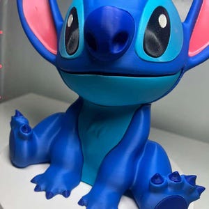 Może przedstawiać: Niebieska figurka postaci Stitch z filmu Disneya Lilo & Stitch. Figurka ma duże czarne oczy, niebieski nos i duże uszy z r&oacute;żowymi wnętrzami. Figurka jest w pozycji siedzącej z wyciągniętymi ramionami i nogami.