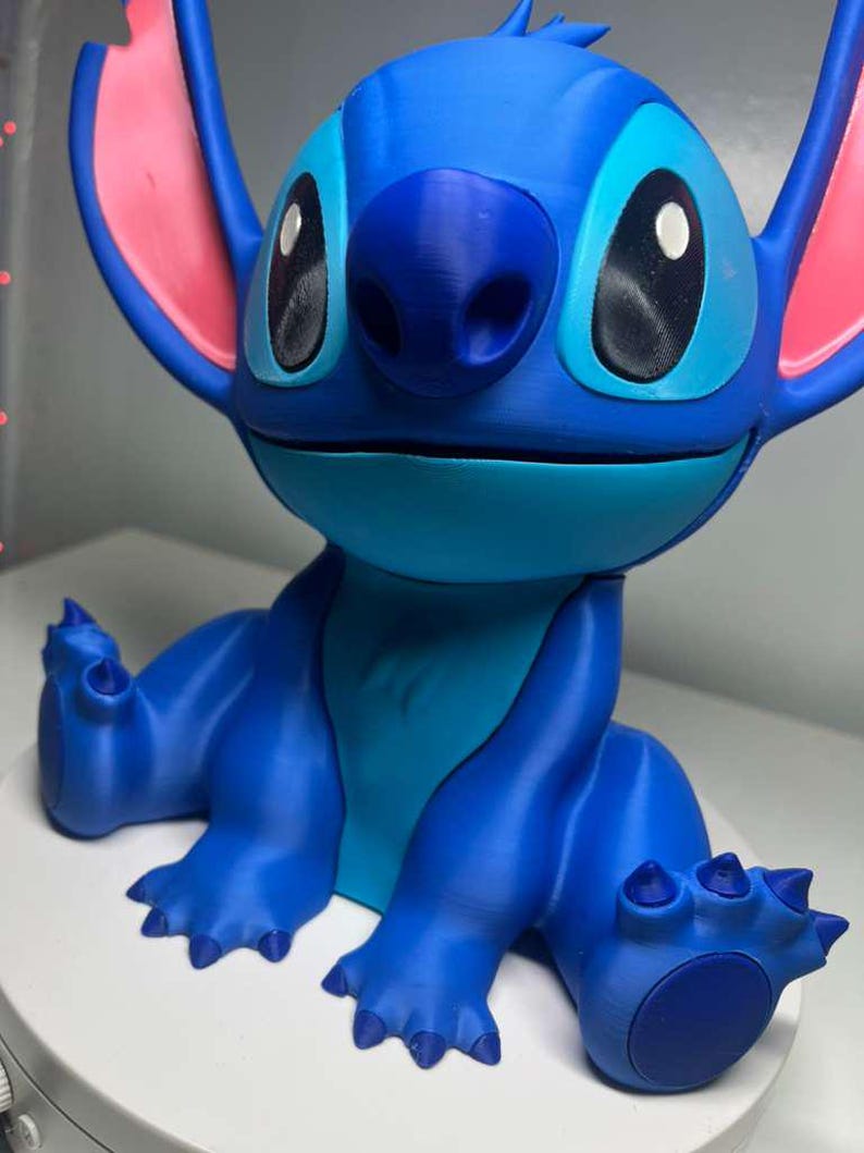 Może przedstawiać: Niebieska figurka postaci Stitch z filmu Disneya Lilo & Stitch. Figurka ma duże czarne oczy, niebieski nos i duże uszy z r&oacute;żowymi wnętrzami. Figurka jest w pozycji siedzącej z wyciągniętymi ramionami i nogami.