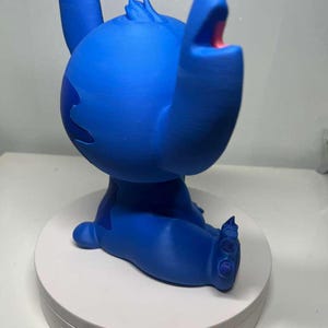 Może przedstawiać: Niebieska figurka Stitcha z Lilo & Stitch, z otwartymi ustami i uniesionymi ramionami. Figurka stoi na białej, okrągłej podstawie.
