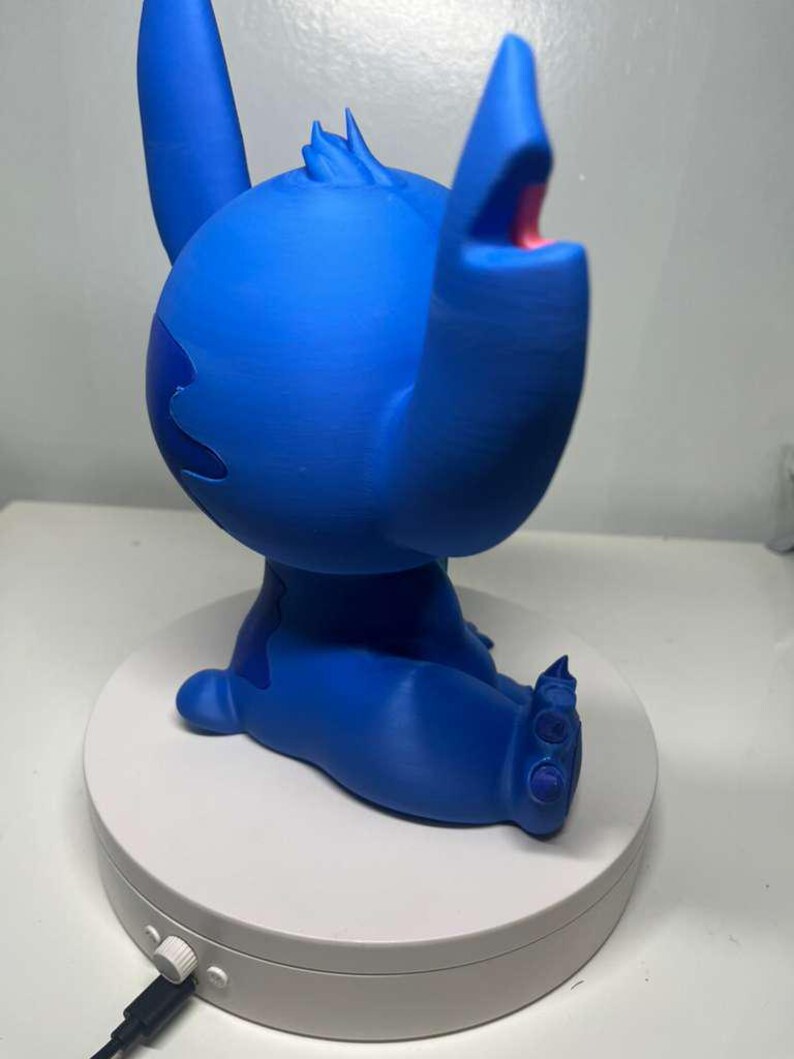 Może przedstawiać: Niebieska figurka Stitcha z Lilo & Stitch, z otwartymi ustami i uniesionymi ramionami. Figurka stoi na białej, okrągłej podstawie.