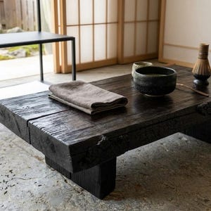 May include: A low, rectangular coffee table made of dark, charred wood with a visible grain and rough, natural edges. On the surface sits a dark ceramic bowl, a bamboo whisk, a small cup, and a folded beige linen cloth. The table has thick legs.
