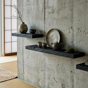 May include: Three black wooden floating shelves mounted on a concrete wall. The shelves hold ceramic pottery, including a teapot, bowls, plates, a vase with a twig, and a yellow candle in a gray holder, arranged in a staggered display.