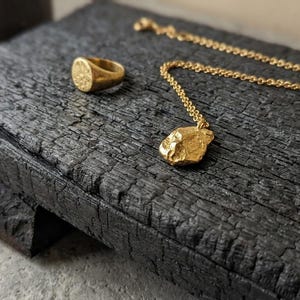 Puede incluir: Un anillo de sello dorado, un collar de cadena dorada con un colgante orgánico texturizado y un brazalete de hilo dorado fino. Las piezas de joyería están dispuestas sobre un bloque de madera carbonizada oscura con una superficie rugosa.