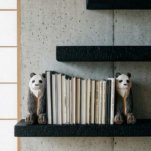 May include: Two carved wooden panda bear bookends with black and white painted details, positioned on a black charred wood shelf holding a row of various books, with two additional empty black shelves mounted on a gray concrete wall.