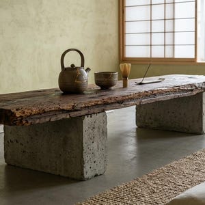 May include: A low rectangular table featuring a thick, dark brown, weathered wooden slab top supported by two large, rectangular, grey concrete blocks. On the table sits a brown ceramic teapot, a matching bowl, a bamboo whisk, and an incense holder.