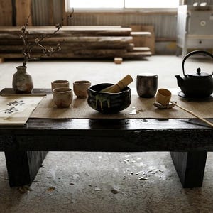 May include: A low, dark wooden table holds a tea set including a black cast iron teapot, a black ceramic bowl with a bamboo whisk, three small beige cups, a small lidded container, a wooden ladle, a small vase with a branch, and a scroll with text.