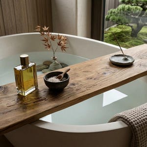 Puede incluir: Una bandeja rectangular de madera para bañera descansa sobre una bañera ovalada blanca. La bandeja contiene una botella de vidrio transparente con líquido amarillo, un cuenco oscuro pequeño con cuchara de madera y un porta-incienso gris.
