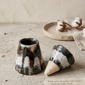 May include: A two-piece ceramic avocado vase with a speckled blue, white, and dark brown drip glaze. The base is cylindrical and the top is conical. A paper note reads: Vase pour avocatier (Awokado) - Japandi Wabi-Sabi. Ø: 9cm, H: 14cm.