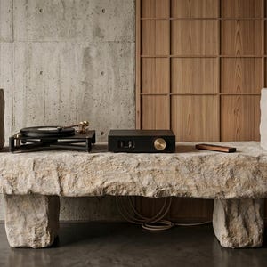 Puede incluir: Un tocadiscos negro, un amplificador negro con un mando dorado y dos altavoces con textura de piedra y rejillas grises. El equipo de audio est&aacute; dispuesto sobre un banco largo y rectangular de piedra toscamente tallada con superficie irregular.