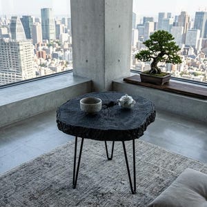 Może przedstawiać: Czarny, okrągły stolik z nogami typu hairpin, mały zestaw do herbaty i drzewko bonsai. Stół wykonany jest z ciemnego drewna i stoi na wzorzystym dywanie. W tle widok na miasto przez duże okno.