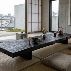 May include: A low, rectangular black wooden table with a live edge sits on a tan tatami mat floor. The table holds a small ceramic vase with flowers, a tea bowl, a whisk, and a copper kettle. Two concrete stools and floor cushions are nearby.