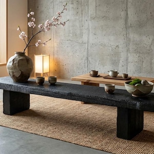 May include: A long, low, rectangular black wooden bench with a charred, textured surface, supported by two thick block legs. On the bench sits a large, round, mottled brown ceramic vase containing a branch with small, pale pink blossoms.