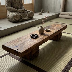 以下が含まれることがあります： 自然な木目調の仕上げが施された長方形の低い木製コーヒーテーブルで、2つのブロック状の脚で支えられています。テーブルの上には、小枝が入った小さなベージュの陶器製花瓶、ダークブラウンのハート型のオブジェ、小さな円形のお香立てが置かれています。