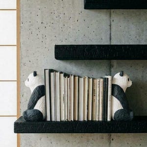 May include: Two black and white panda-shaped bookends positioned on a black textured shelf, holding a row of books upright. The panda figures are facing inward toward the books, which have various light-colored spines and are arranged in a vertical row.