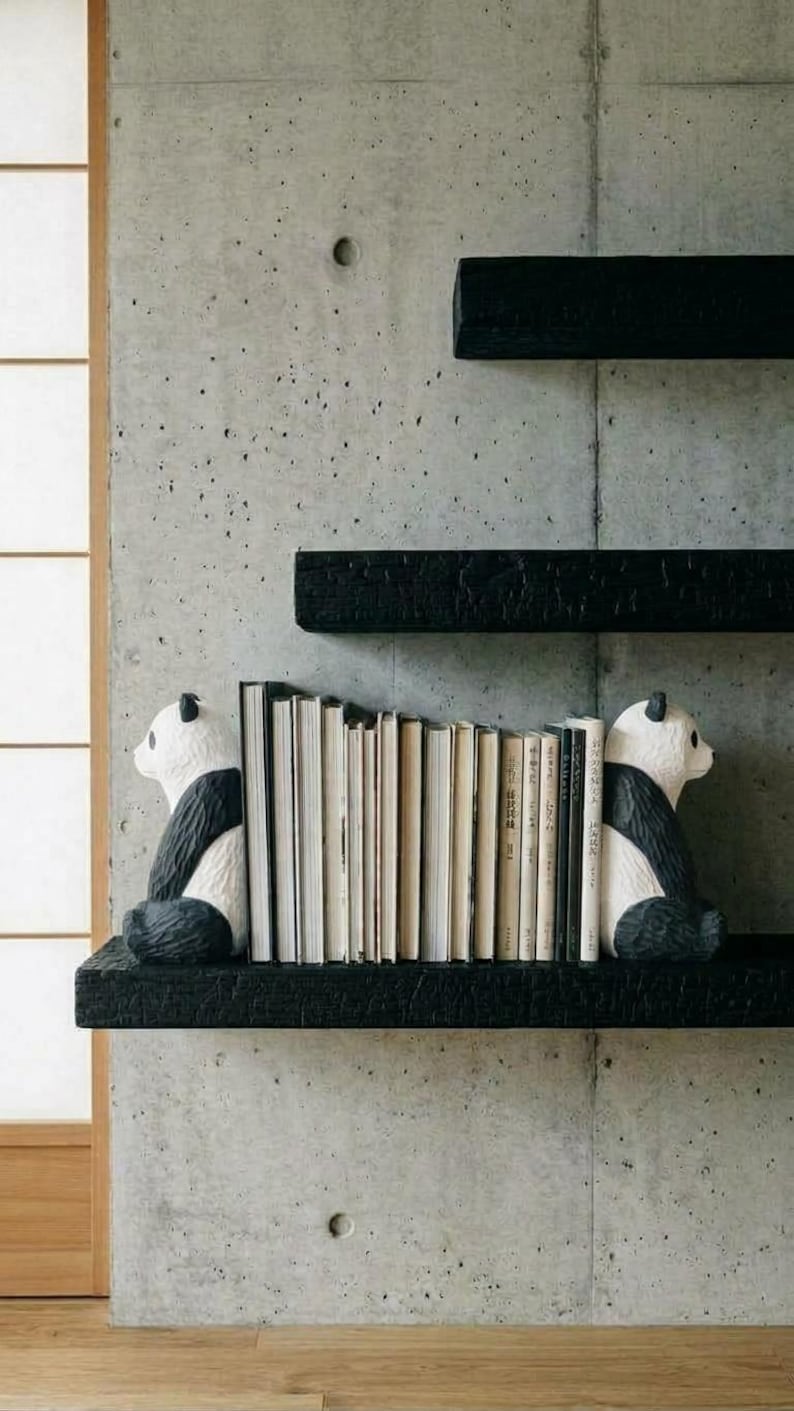 May include: Two black and white panda-shaped bookends positioned on a black textured shelf, holding a row of books upright. The panda figures are facing inward toward the books, which have various light-colored spines and are arranged in a vertical row.