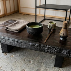 Könnte beinhalten: Ein niedriger, rechteckiger Couchtisch aus dunklem, verkohltem Holz mit natürlicher Baumkante. Auf dem Tisch liegen ein gefaltetes beiges Leinentuch, eine schwarze Keramikschale mit grünem Matcha-Tee, eine kleine Keramiktasse, ein Holzlöffel und ein Bambusbesen.
