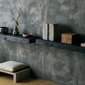 May include: Two black charred wood floating shelves mounted on a gray textured wall. The left shelf holds a dark bowl, a small bare branch, and a dish. The right shelf holds a gray block, a wooden box with text, and two stones on a book.