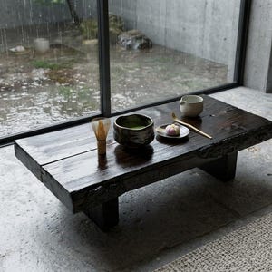 May include: A low, dark-stained wooden coffee table with a live edge, featuring a bamboo whisk, a dark ceramic bowl filled with green matcha tea, a small white plate with a pink confection, and a bamboo tea scoop arranged on its surface.