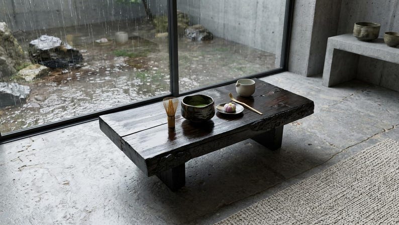 May include: A low, dark-stained wooden coffee table with a live edge, featuring a bamboo whisk, a dark ceramic bowl filled with green matcha tea, a small white plate with a pink confection, and a bamboo tea scoop arranged on its surface.