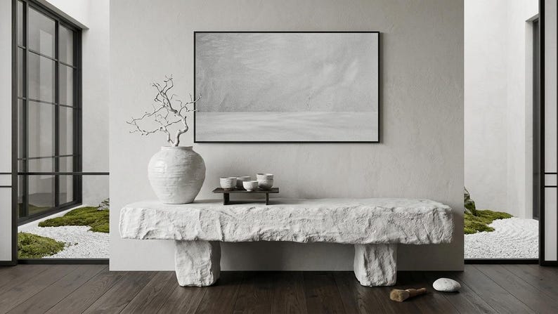 Puede incluir: Un banco de piedra rectangular y largo con una superficie rugosa y texturizada, sostenido por dos patas en bloque. Encima hay un jarr&oacute;n de cer&aacute;mica blanca grande y redondo con una rama desnuda y retorcida, junto a una bandeja oscura peque&ntilde;a.
