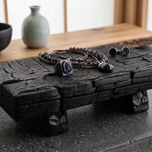 May include: A rectangular, charred black wooden display stand with four short legs. Resting on the textured, cracked surface are a silver ring with a dark blue stone, a dark beaded necklace, and a pair of small, round silver cufflinks.
