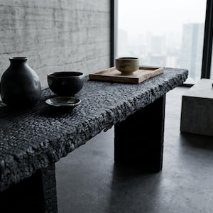 Op de afbeelding: Een minimalistische interieurscène met een donkere, getextureerde houten tafel met een vaas, kommen en een klein bord. Een houten dienblad met een kom staat op de tafel. Een betonnen bank en een tak zijn ook in de kamer aanwezig.