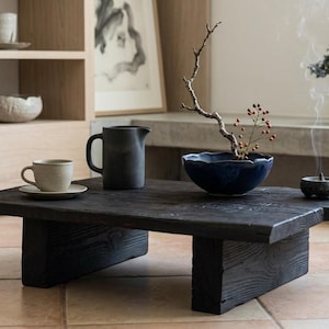 May include: A low, rectangular coffee table made of dark, charred wood with a thick, block-style base. On the table sits a light-colored ceramic cup and saucer, a matte black pitcher, a deep blue bowl with a thin branch, and a small incense burner.