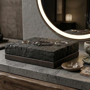 May include: A square, charred wood jewellery display stand with a dark wooden base featuring the engraved name HARRIET. The top surface is textured and black, holding three pieces of jewellery including a ring, a bracelet, and a thin chain.