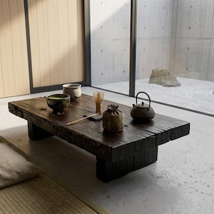 May include: A low, dark-stained wooden table with a rough, textured edge sits on a concrete floor. On the table are a green tea bowl, a small ceramic cup, a bamboo whisk, a small brown pouch, and a dark metal teapot with a curved handle.