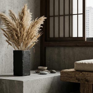 Puede incluir: Un jarrón negro rectangular con superficie texturizada contiene un manojo de hierba de la pampa beige seca. El jarrón está sobre una repisa de hormigón, con un banco de madera y una ventana al fondo. La escena evoca una estética minimalista y natural.