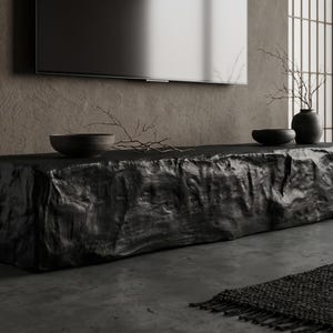 May include: A long, rectangular black media console with a rough, textured, rock-like surface. The console features a flat top surface holding two dark bowls and a small branch, positioned below a wall-mounted television screen.
