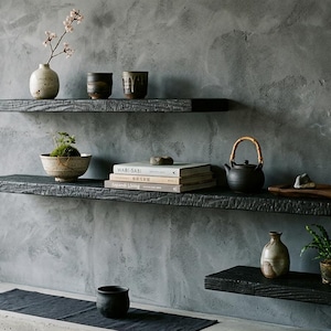 May include: Three black floating shelves with a charred wood texture mounted on a gray wall. The shelves hold various items including a ceramic vase with a branch, small bowls, a stack of books, a dark teapot, a wooden tray, and small plants.