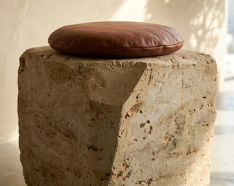 May include: A cube-shaped stool made of rough, porous beige stone with a smooth, circular brown leather cushion placed on top. The stone base features natural textures and uneven edges, providing a contrast to the soft, rounded leather seat.