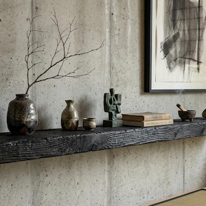 May include: A long, rectangular, charred black wooden floating shelf mounted on a concrete wall. The shelf holds a dark ceramic vase with a bare branch, two smaller ceramic vessels, a green abstract sculpture, two stacked books, and a small bowl.