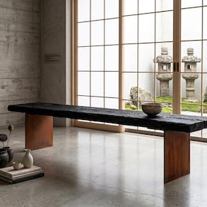 May include: A long bench featuring a thick, charred black wooden seat supported by two vertical, rust-colored metal slab legs. A single ceramic bowl rests on the bench surface, while several dark pottery vessels and a stack of books sit nearby.