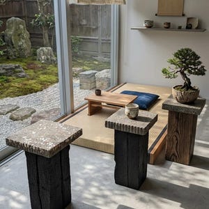 Puede incluir: Dos pedestales cuadrados con tapa de piedra y base de madera negra carbonizada sobre un suelo de hormigón. Cada pedestal presenta una superficie de piedra texturizada y moteada. Una pequeña taza de cerámica descansa sobre uno, un bonsái sobre el otro.