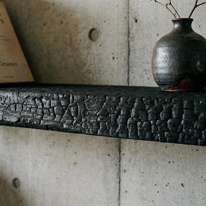 May include: A horizontal wall shelf made of charred black wood with a deeply textured, crackled surface. The shelf holds a dark brown ceramic vase containing thin twigs, a light-colored book titled Zen Mind, Beginner's Mind, and a rock.