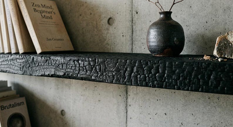 May include: A horizontal wall shelf made of charred black wood with a deeply textured, crackled surface. The shelf holds a dark brown ceramic vase containing thin twigs, a light-colored book titled Zen Mind, Beginner's Mind, and a rock.