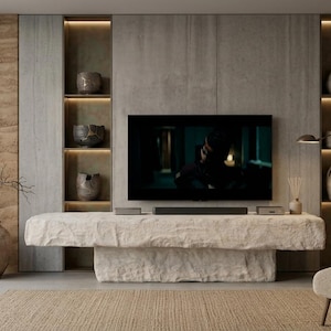 Pode incluir: Uma sala de estar com uma grande consola multimédia de pedra texturizada em branco sujo, centrada sob uma televisão de parede. A consola é ladeada por duas estantes verticais iluminadas com vários vasos de cerâmica e objetos.